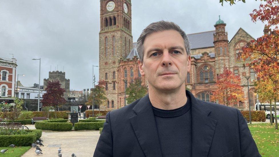 Shaun Harkin stands in Derry's peace garden. The city's Guildhall can be seen in the background as can the tower museum. He is wearing a black jacket over a back jumper. 