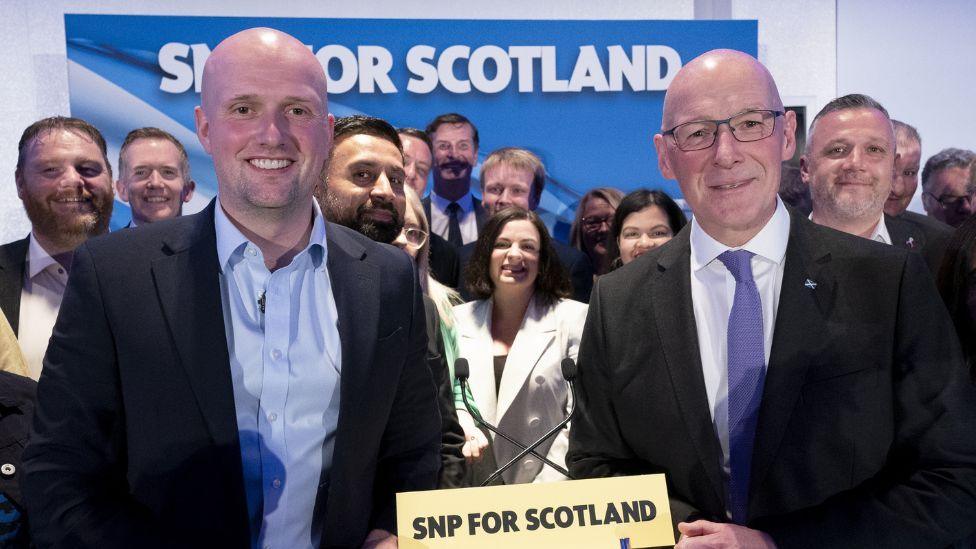 Stephen Flynn, a bald man wearing a dark suit and blue shirt, and John Swinney, a bald man wearing glasses and a dark suit with a white shirt and purple tie, smile at the camera in front of SNP colleagues at a party press conference 