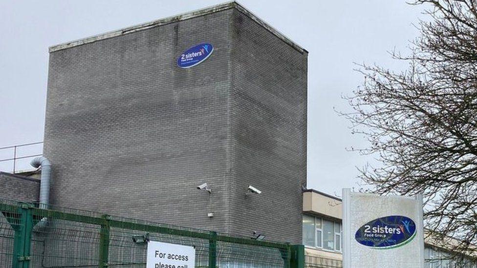 A grey building with a blue oval logo that reads 2 sisters. There is a white sign in the right foreground that also has the same logo.
