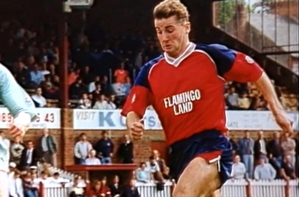 David Longhurst playing a game wearing football kit that is red and blue and says "Flamingo Land"