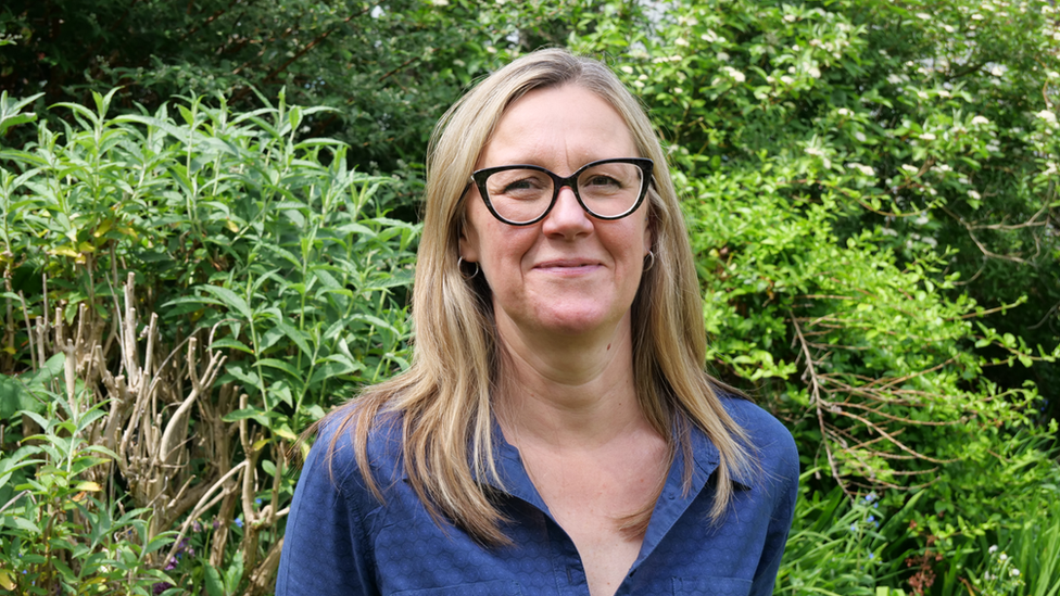 Debbie Tann stands in front of bushes and trees. She has long, fair hair and wears glasses and a dark blue shirt.