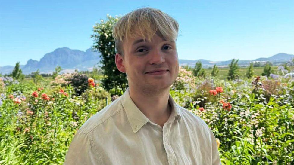 A photo of Erik Ibsen smiling at the camera. He has blonde hair that comes down to just above his eyes and is wearing a very light coloured yellow shirt that is buttoned up. Behind him is a picturesque scene of green bushes and general greenery with some mountains in the far background.
