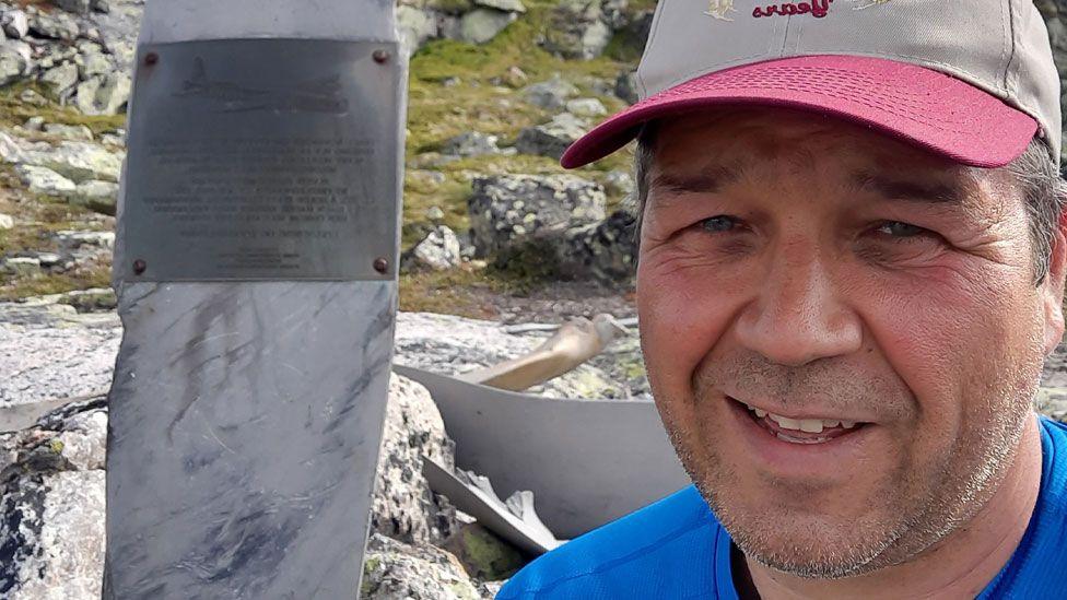 Espen Thronsen, wearing a bright blue T-shirt and beige and pink baseball cap, on a rocky Norwegian mountainside. He is on the right and starting to smile. On the left is a close-up of a war memorial made out of an aeroplane propeller. There is a grey metal plaque with writing attached to it.