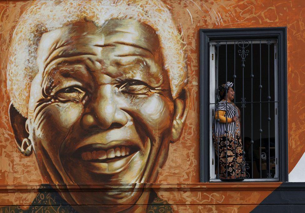 A large sepia-toned mural of a smiling Nelson Mandela covers the side of a building. A woman stands and smiles in one of the windows as she poses for a photo.