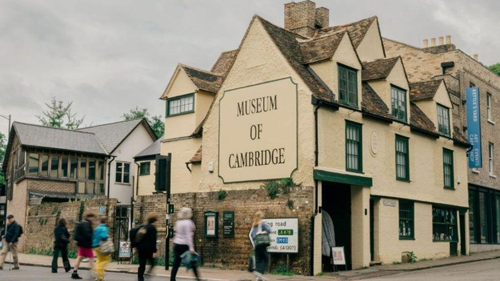 The Museum of Cambridge, a cream-painted three storey 16th century former coaching inn, which has green-painted windows. The image shows the museum at right angles to a junction. Its gable end has Museum of Cambridge written in black letters. People are crossing the road towards it.