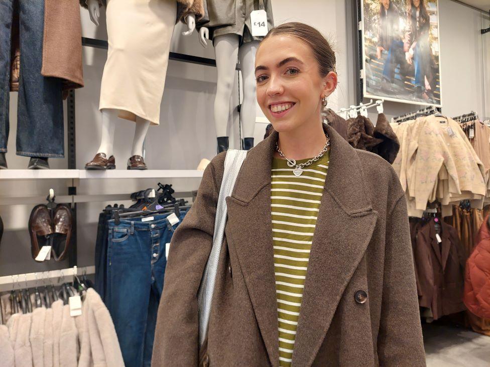 A woman with her hair tied up, in a yellow striped top and brown coat, smiles at the camera as she stands inside a Primark store
