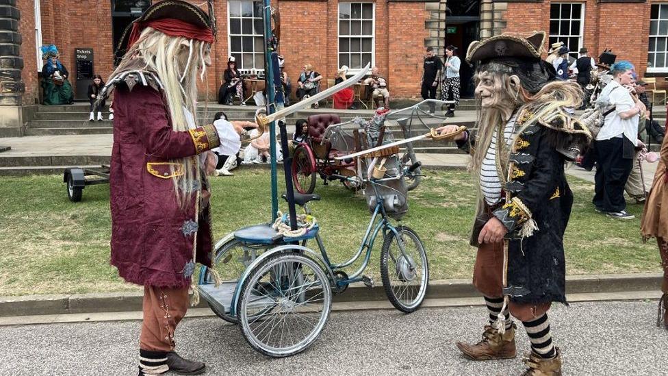 Two people stand facing each other with long wooden sticks. They are dressed as pirates but have face masks on and are stood next to an old style bike, other people are seen in the background milling about. 