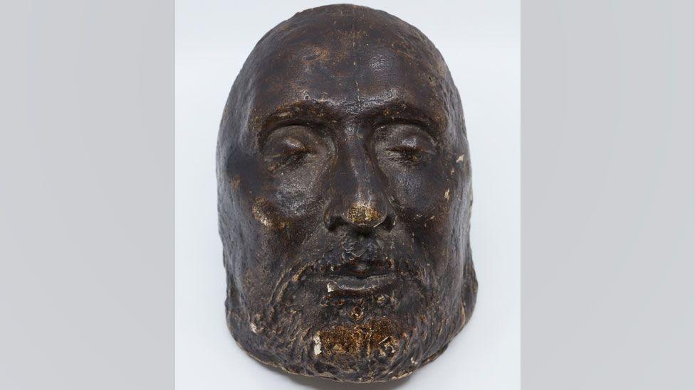 A funeral mask for Oliver Cromwell. It shows his bearded face. His eyes are closed. It is made from a very dark material