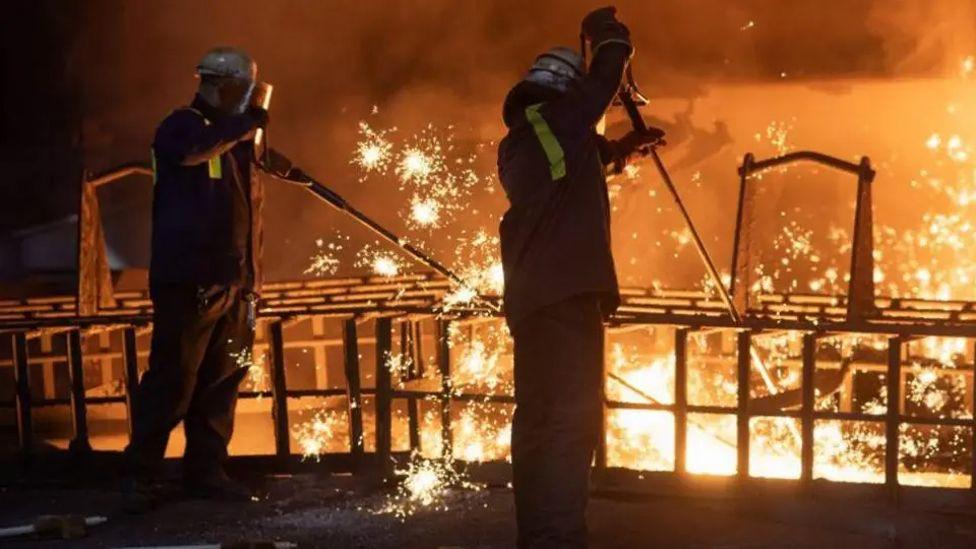 Two steelworkers in safety jackets, visors and helmets stand in a dark room near orange molten metal, with bright sparks around them.