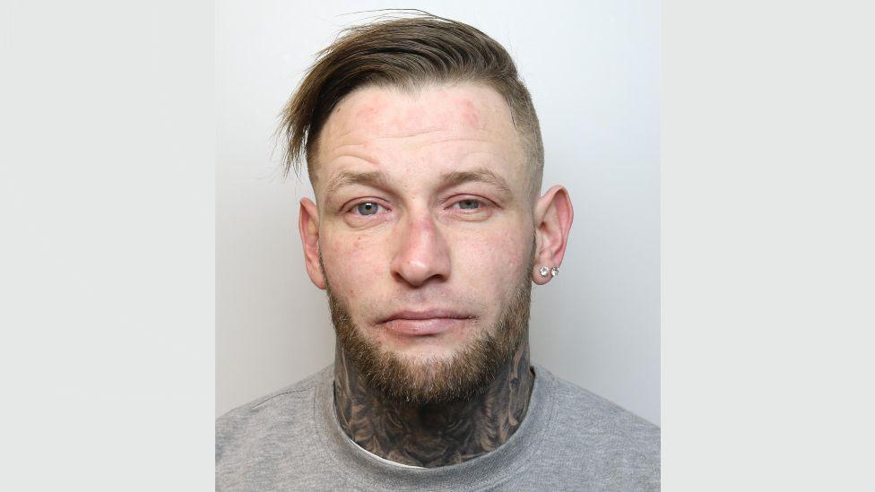 A police mugshot of Casey Delaney. He is wearing a grey t-shirt and standing against a grey background. His brown hair swept to the right on top with the sides shaved. He has blue eyes, a brown trimmed beard with no moustache and two diamond earrings in his left earlobe. He is looking at the camera with a blank expression.