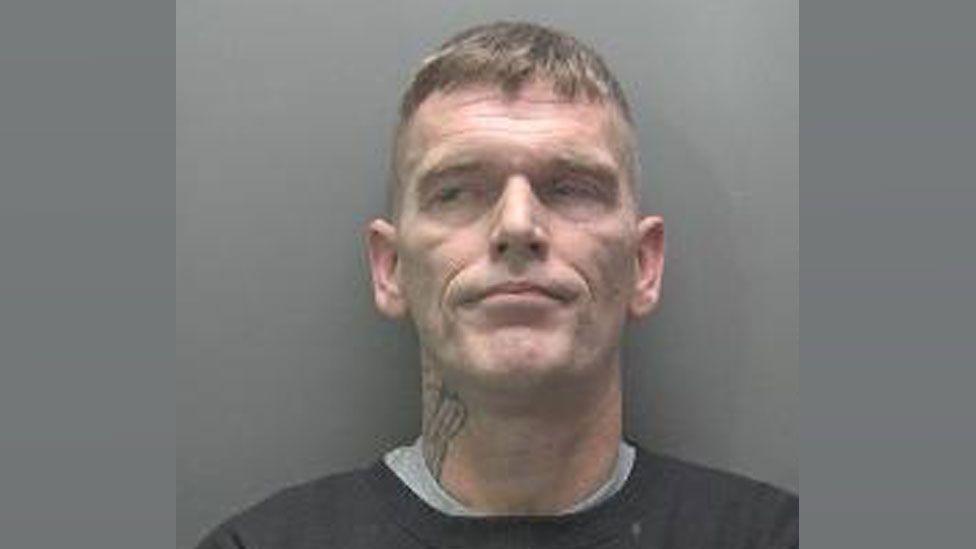 Andrew Jenkins in a custody photo. He is clean shaven and has cropped hair on his sides and longer light brown hair on top. He is wearing a black sweatshirt over a grey t-shirt and has a tattoo on the right side of his neck.