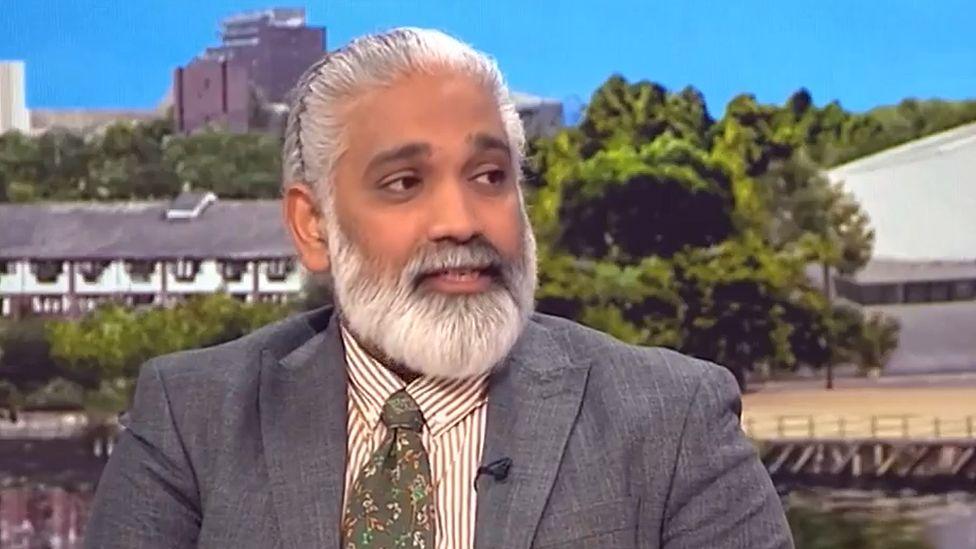 Dr Sakthi Karunanithi in a TV studio in front of a screen showing a row of cottages and some trees under a cloudless blue sky. He is looking to the right and has long, neat white hair tied back and a beard and is wearing a grey suit with a white and brown striped shirt and floral green tie