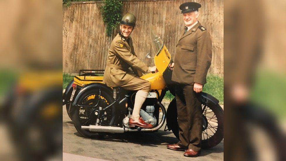 An old, faded photograph of Derek seated on a vintage yellow AA motorbike with a fellow patrolman standing next to him. Both are dressed in the uniform of the time - khaki-coloured jackets and trousers.