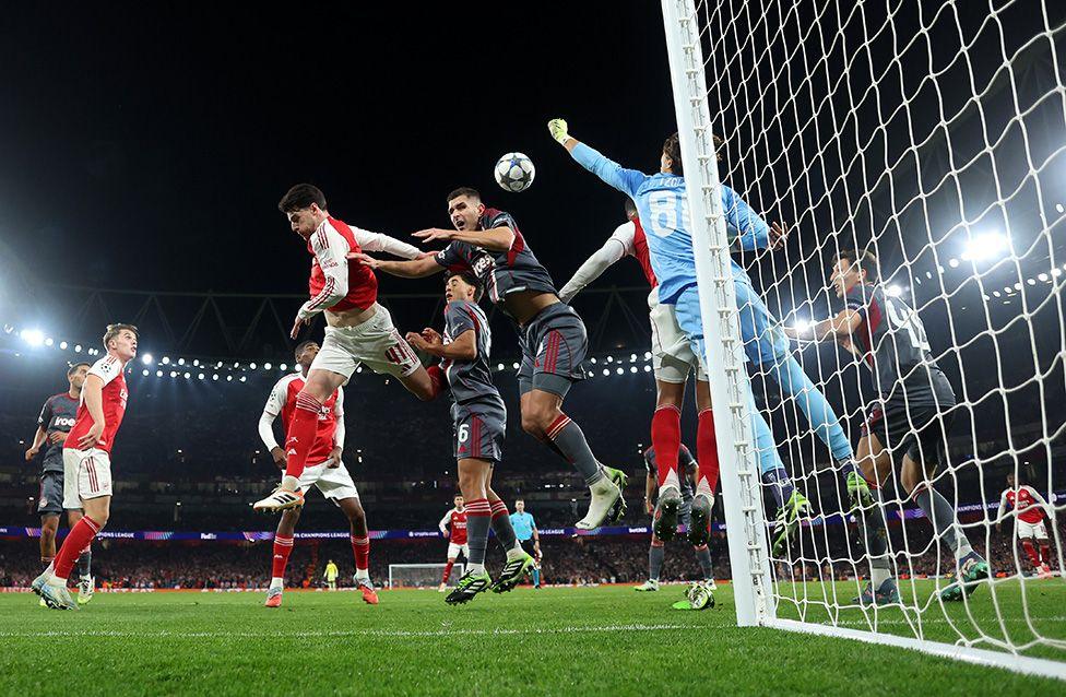 Arsenal’s Declan Rice attempts a header as Olympiacos goalkeeper Kostas Tzolakis punches the ball clear during a Uefa Champions League match in London. The action takes place in front of the goal, with multiple players contesting under bright stadium lights.