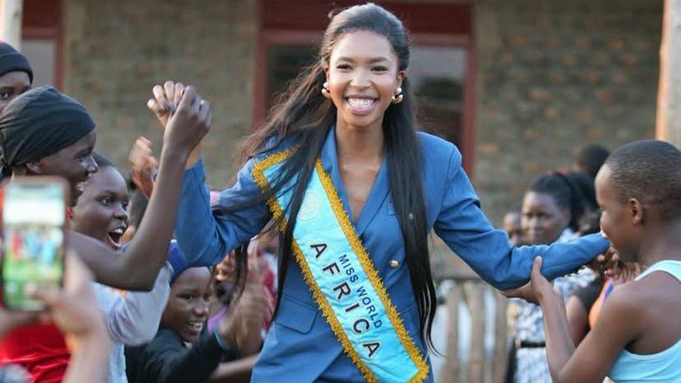 Lesego Chombo in a blue suit with a Miss World Africa sash bhold the hands of young girls