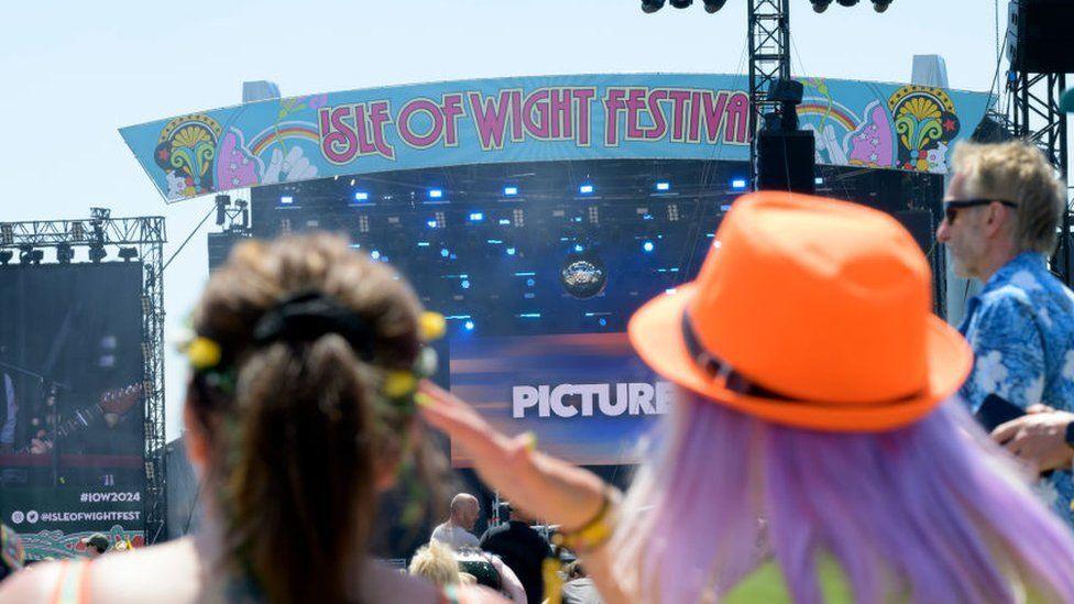 Isle of Wight Festival's main stage from behind two festival goers, one with long purple hair and a bright orange hat, and the other with dark hair tied back in a ponytail