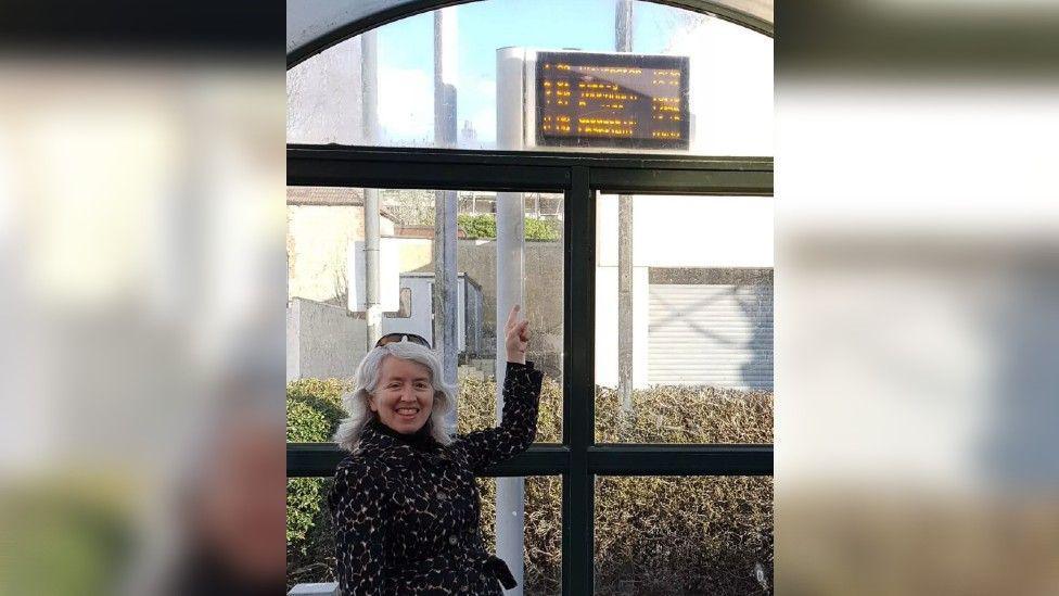 Wendy in a bus stop pointing to the arrivals board above her