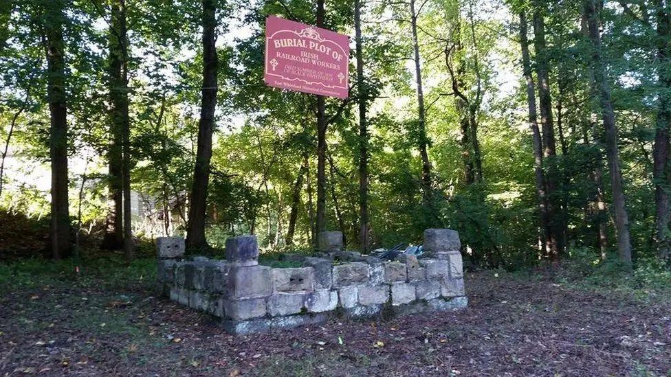 The picture shows a stone memorial in a forest. A red sign on the memorial reads: "Burial plot of Irish railroad workers".