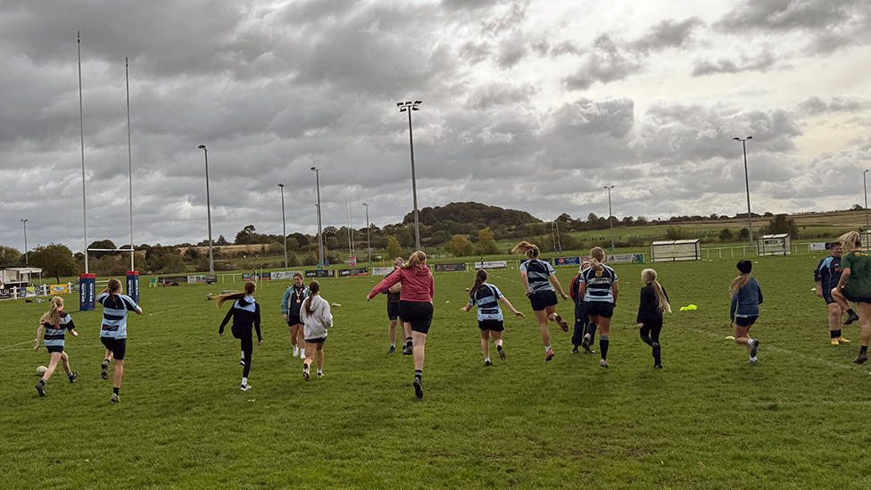 A green rugby pitch with a line of young girls running with their backs to the camera. They are wearing shorts and white and navy rugby tops