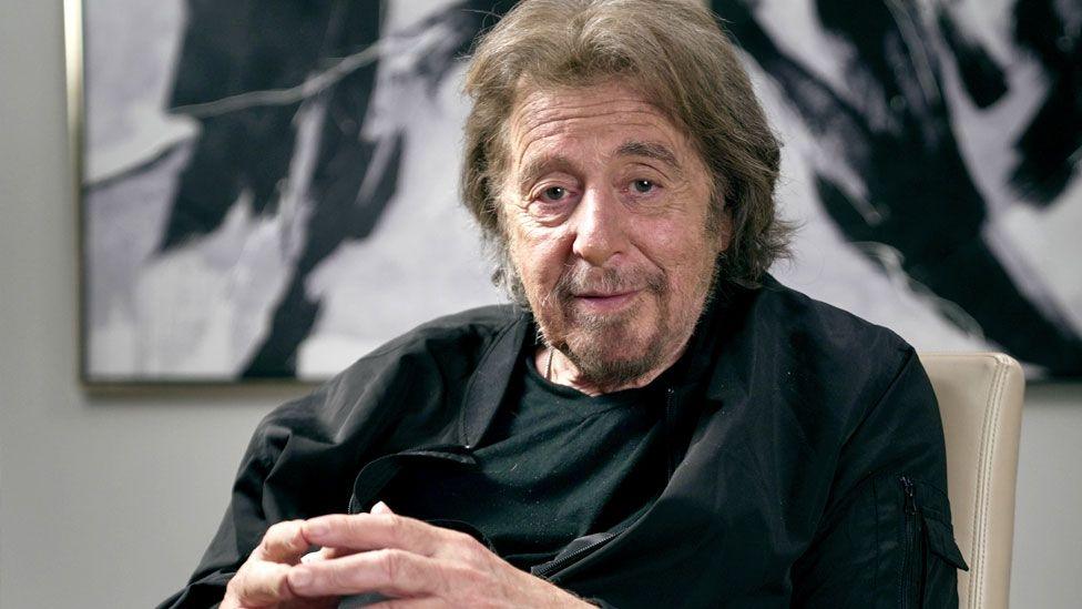 Al Pacino sits and looks directly at the camera, his fingertips touching, with a piece of art behind him on the wall