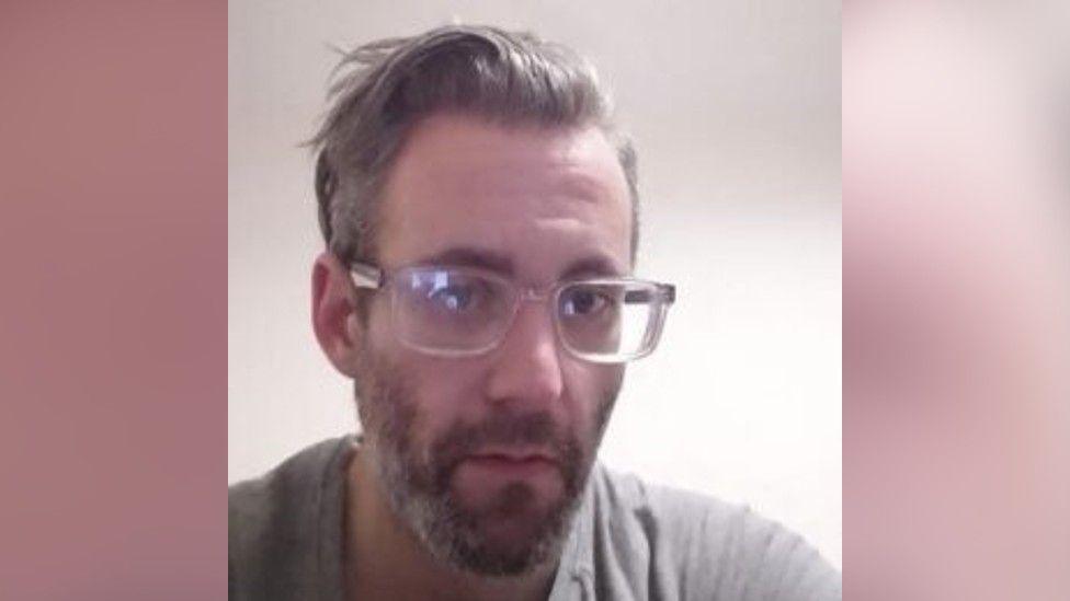 A close-up image of Michael Wheeler. He has grey hair, longer on top and swept back, and wears clear-framed square glasses. 