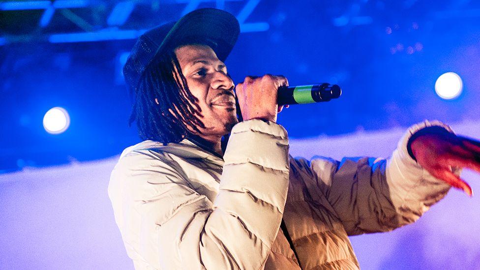 MC Bushkin of Heartless Crew performs onstage, wearing a dark coloured cap, cream coloured puffer jacket with a black microphone in hand.