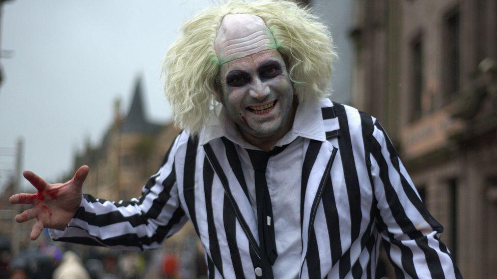 A man dressed as Beetlejuice smiles directly into the camera at the Paisley Halloween Festival. He is wearing white face paint, with dark face paint around his eyes. His clothing is a white shirt with a black tie under a black and white striped jacket. He has a wig on his head which is bald in the middle with green paint on the sides and light green hair coming out the top. His right hand has red marks on it.