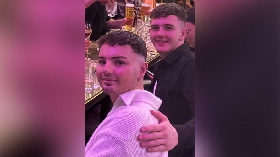 Two young men, dressed in smart-casual clothing, sit next to each other at a long table dotted with empty drinks glasses. Both are looking over their left shoulder at the camera, and the young man at the rear of the shot has his hand resting affectionately on the other young man's shoulder