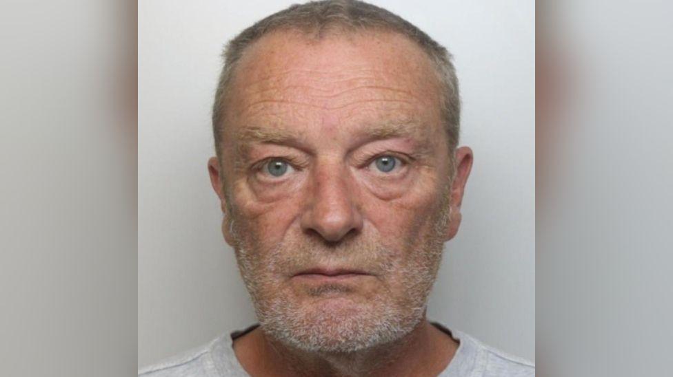 Paul Renwick looking at the camera in a police mugshot. He is an older man with short, graying hair and a beard or stubble. 