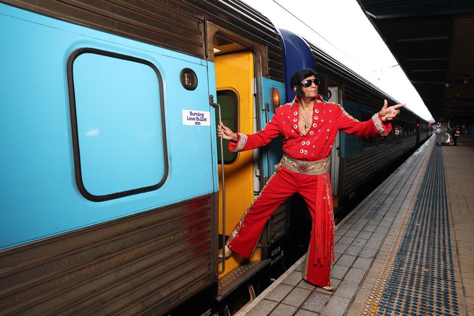 An Elvis impersonator wearing in a red jumpsuit points to the sky as he steps on to a train.