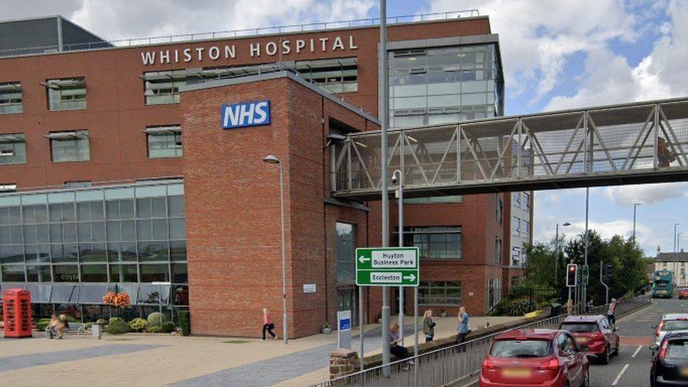 Whiston Hospital - a red brick building - with a walkway over the road which has traffic on it 