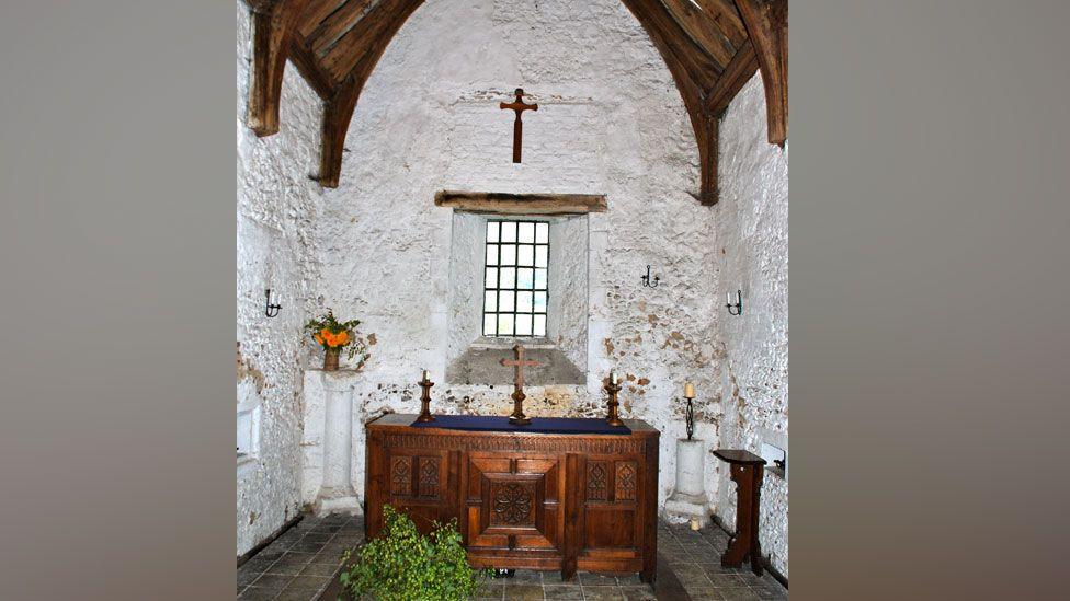 Interior of Cambridge Leper Chapel, showing white washed walls, a wooden beamed roof above, a wooden altar below a window, with a cross hanging on the wall above it