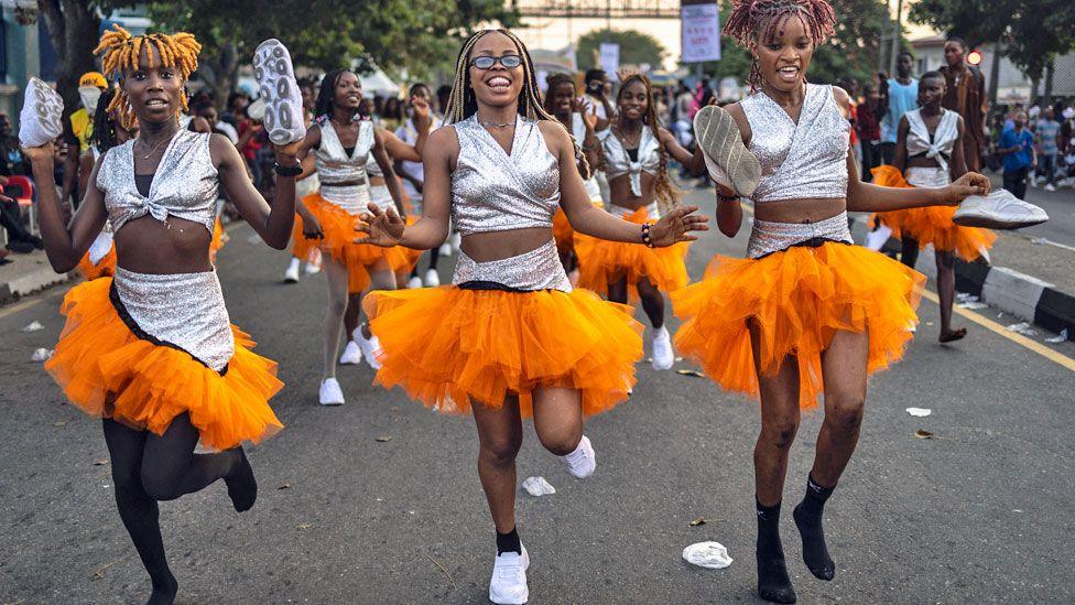 Nigeria dancers in silver tops and orange pom pom skirts skip down a street at Calabar's carnival