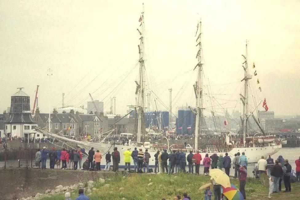 White tall ship with three masts, with Aberdeen in the background and spectators in the foreground.