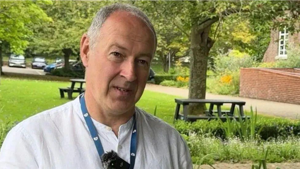 A man wearing a white shirt and blue lanyard stood in a green area with dark green benches. 