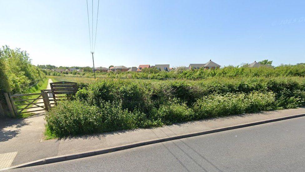 Road and path with shrubs and hedging wooden gate and sign into field with path winding along to the left - blue sky, sunny day.