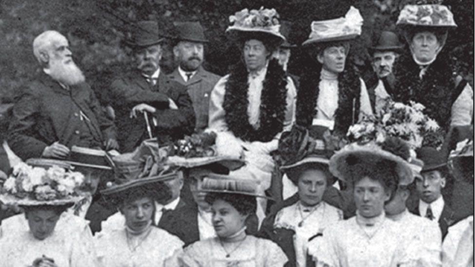 A black and white Edwardian photo of a group of men and women. Allan Minns is in the back row, second left, with folded arms, wearing a hat, holding a stick. He has a drooping large moustache covering his upper lip. Ophelia Minns is the first woman on the right on the back row. She is wearing a white dress, has a floral garland around her neck and is wearing a large flower-covered hat