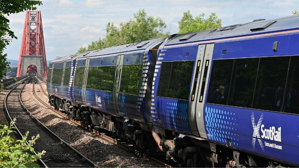 A ScotRail trains heads along the tracks towards the Forth Rail Bridge on a sunny day.