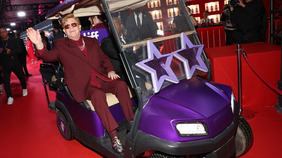 Sir Elton waving and riding in a golf cart-type buggy