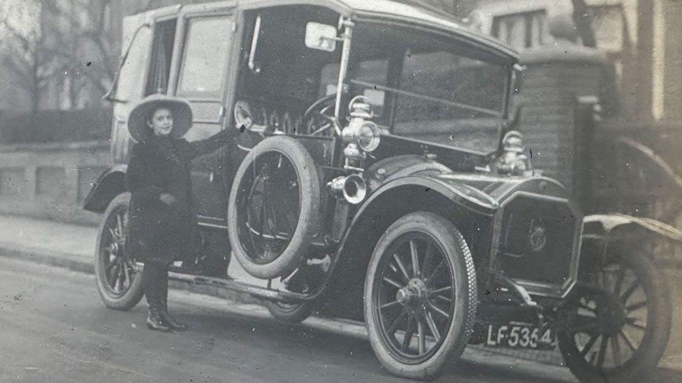 A black and white mage showing Elsa beside a car on Woodborough Road, Putney, London. She is wearing a broad brim hat, a black knee-length coat over black tights and shoes. Her left hand is holding the window rim of the car on the driver's side and she is smiling.