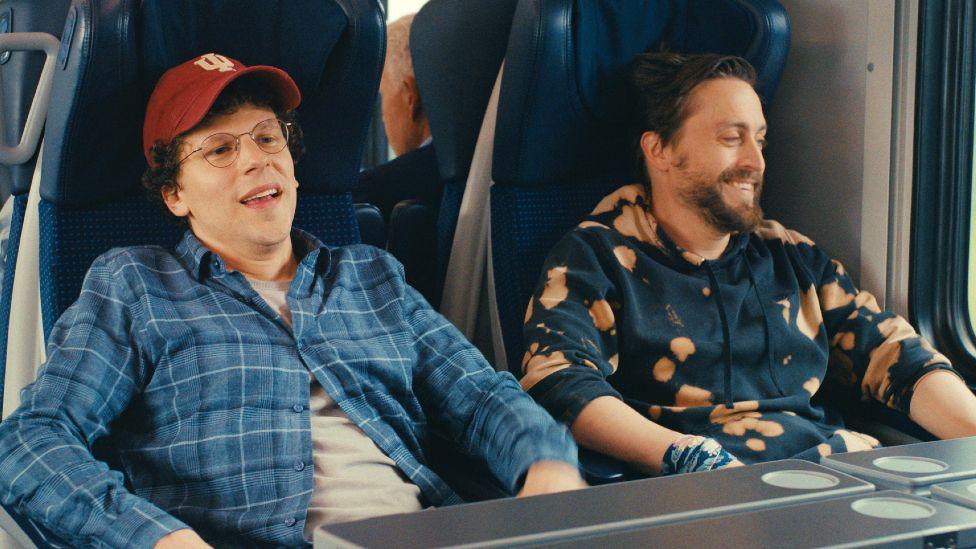 Jesse Eisenberg is wearing a checked blue shirt and grey t-shirt and Kieran Culkin is wearing a navy and peach hoody, they are laughing while sitting on a train