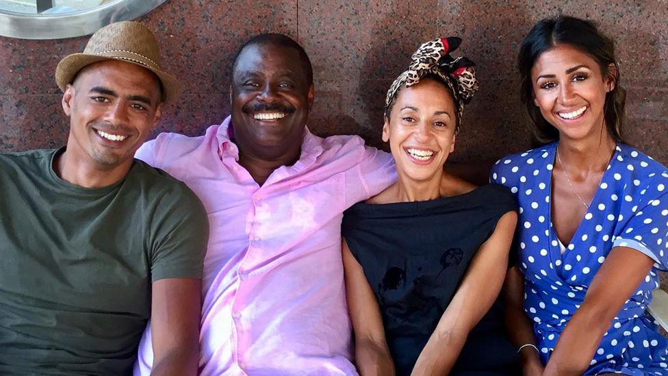 Mel Tottoh with his three children, a son and two daughters, all grown up, smiling and wearing casual clothes as they sit beside their father who is wearing a pink shirt