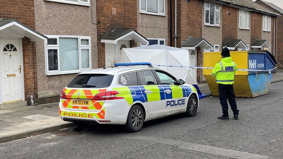 A police officer and parked police car next to a terraced house which has been cordoned off. A white tent is in front of the door area. There is also a yellow and blue metal skip to the right of the image.