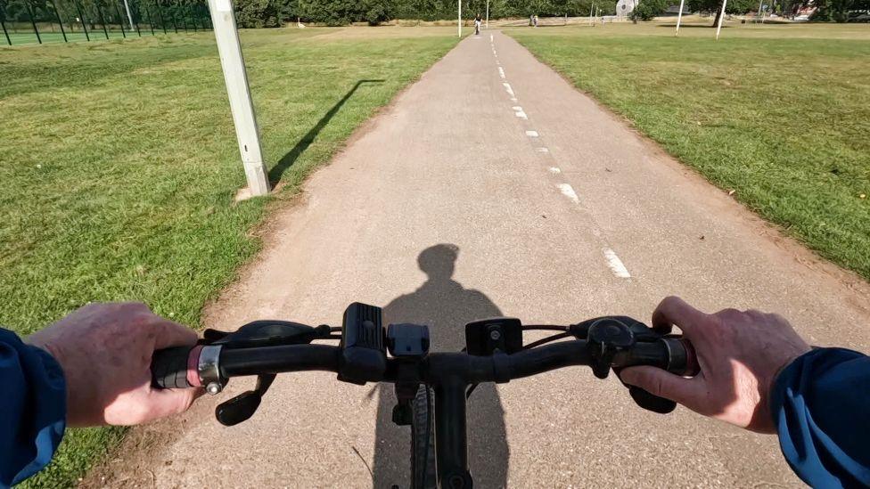A first-person view of someone riding a bicycle along a paved path through a flat grassy landscape in a park. The rider’s hands can be seen on the handlebars, and their shadow stretches ahead on the path. The path itself has dashed white lines down the middle.