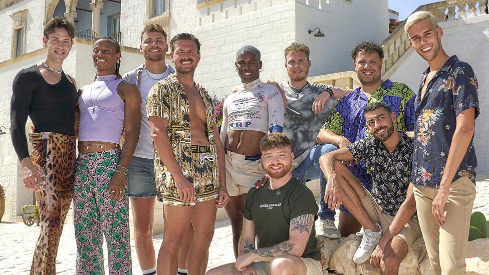 A group of ten men, stood outside a white villa in the sunshine. They are all wearing colourful clothing, and many have their arms round each other as they smile at the camera.