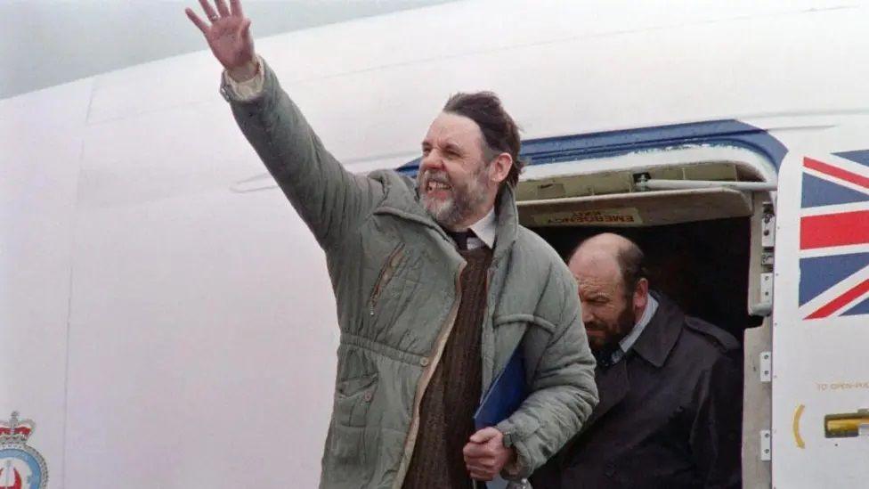 SirTerry Waite with medium-length dark hair and beard. He is wearing a green coat over a brown sweater and brown tie. He is waving with his right hand while standing on the top step of passenger stairs attached to an aircraft which has a union flag painted next to the door. There is another man with a beard behind Sir Terry.