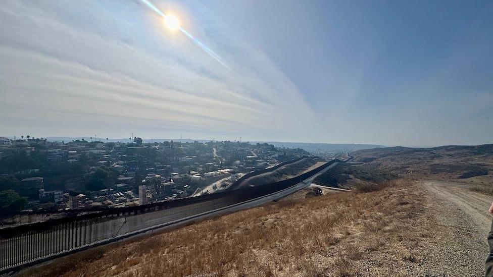 The US-Mexico border shows one side is dry brush, the other, an urban landscape