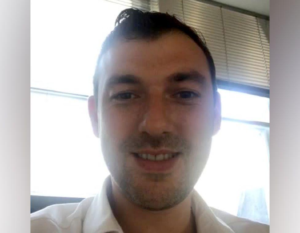 Gregory Assemakis: A man dark hair and stubble wearing an open-necked light shirt.