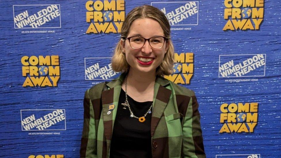 Lydia Greatrix smiles and looks directly at the camera as she stands in front of a billboard for Come From Away. She is wearing a green plaid jacket and black top, with a necklace with a sunflower on it. She has short blonde hair and glasses. 