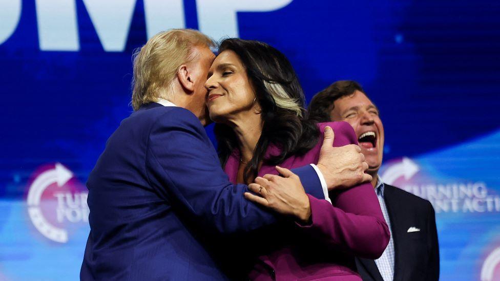 Trump and Gabbard embrace on stage in Duluth, Georgia, on campaign trail in October, with Tucker Carlson in background laughing